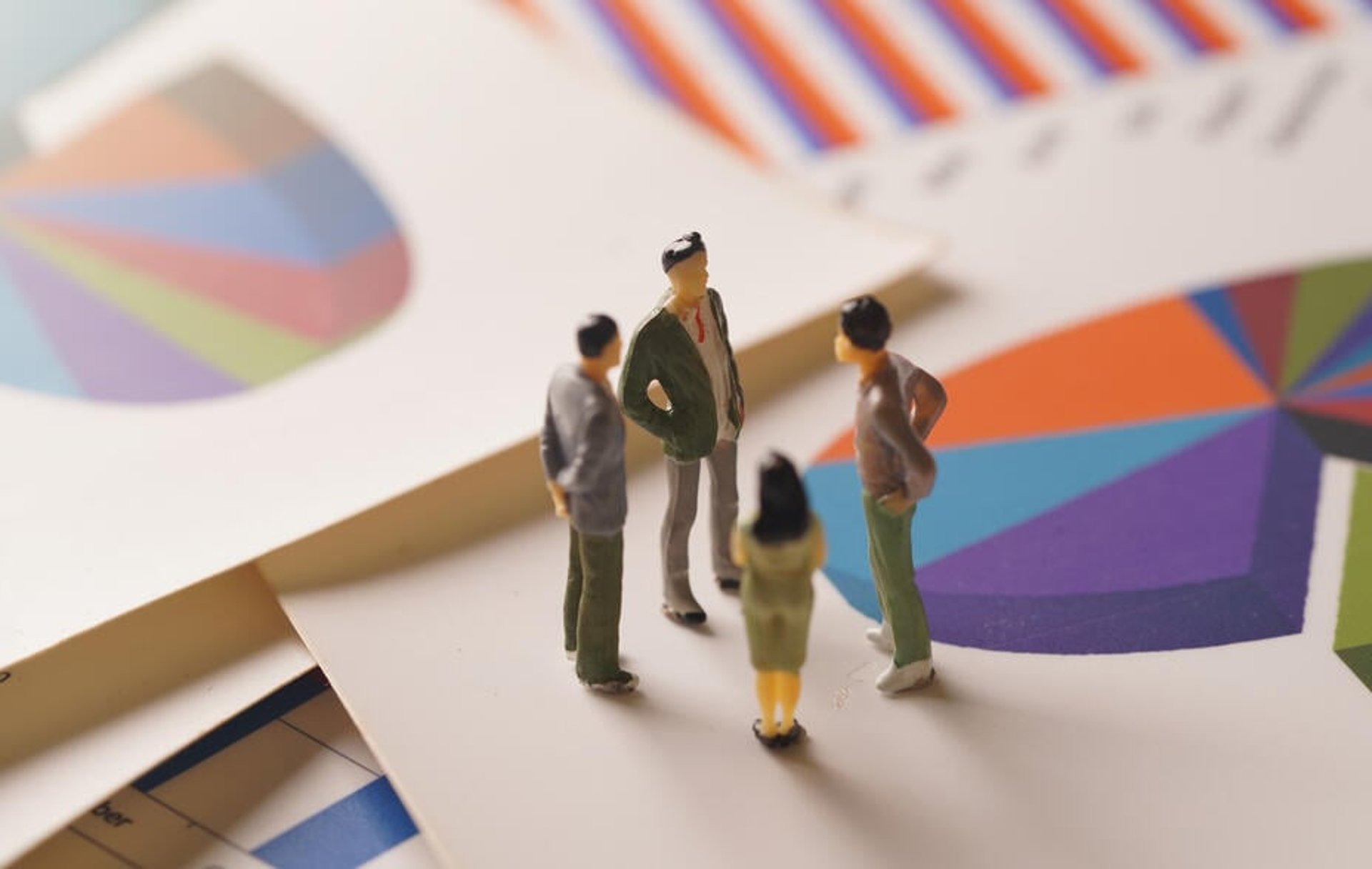 Miniature figurines of businesspeople are posed in a group on top of colourful print-outs of charts and graphs