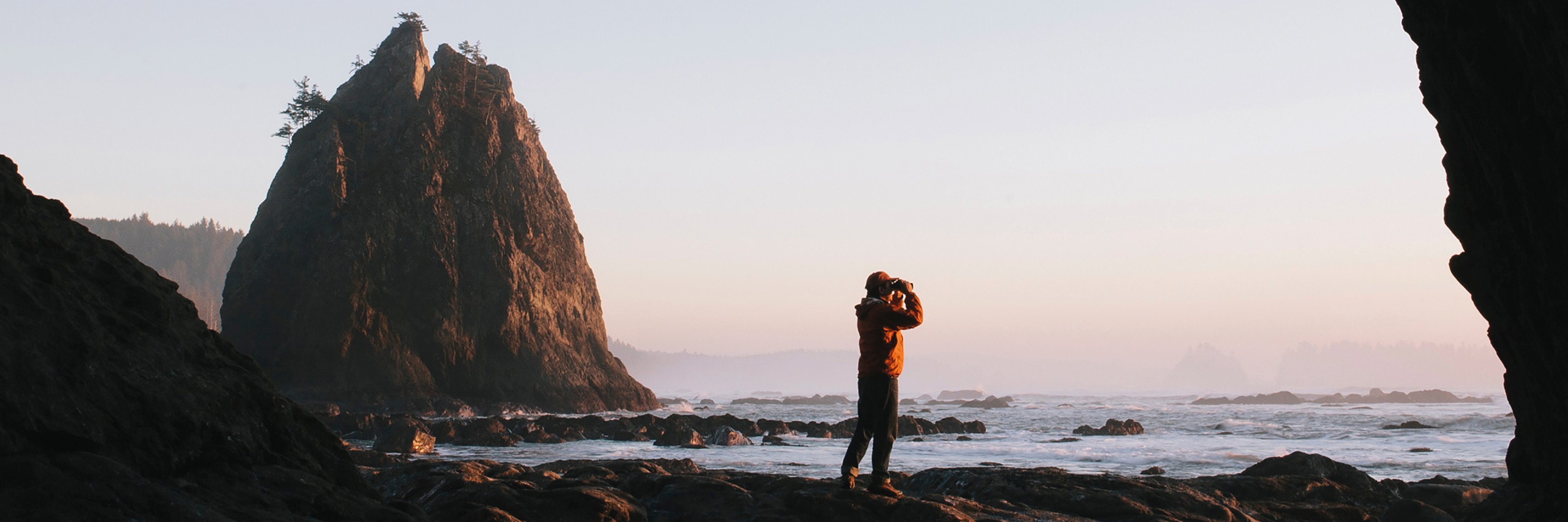 A person standing on a beach beneath sea cave at dusk, with the sea and coastline beyond, using binoculars at Hole-in-the-Rock, Rialto Beach