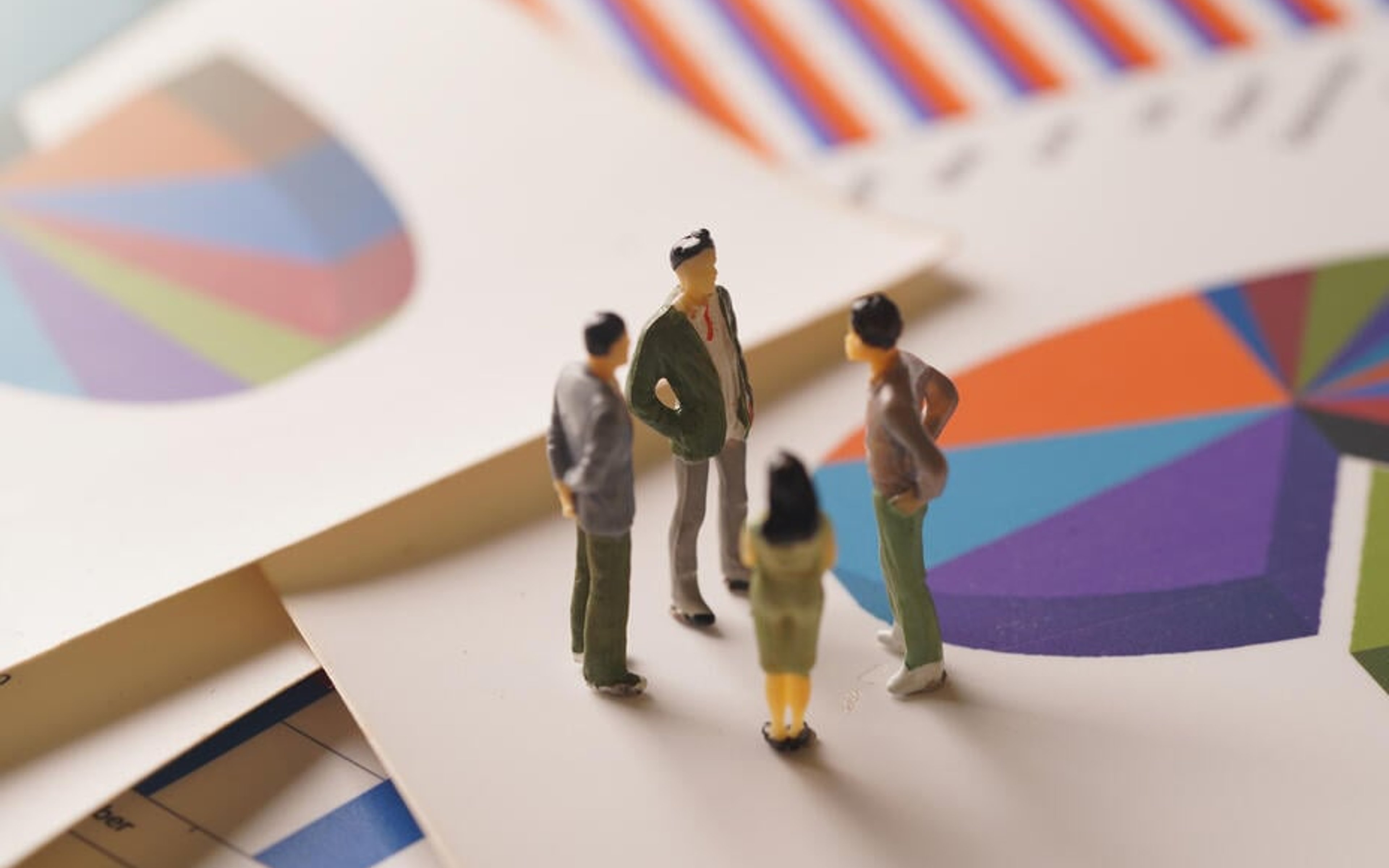 Miniature figurines of businesspeople are posed in a group on top of colourful print-outs of charts and graphs