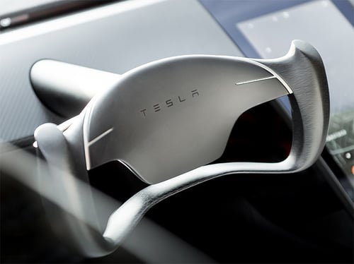 A close-up of a Tesla yoke steering wheel inside a vehicle. The steering wheel features a minimalist design with smooth curves and the Tesla logo in the center.